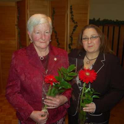 Two women are holding flowers, smiling to the camera, and hugging