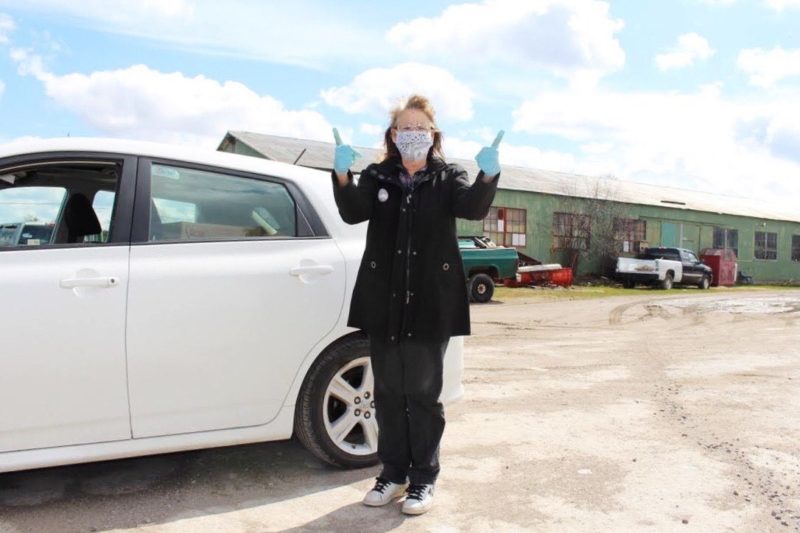 A picture of a woman standing in front of a car, wearing a mask, and giving two thumbs up