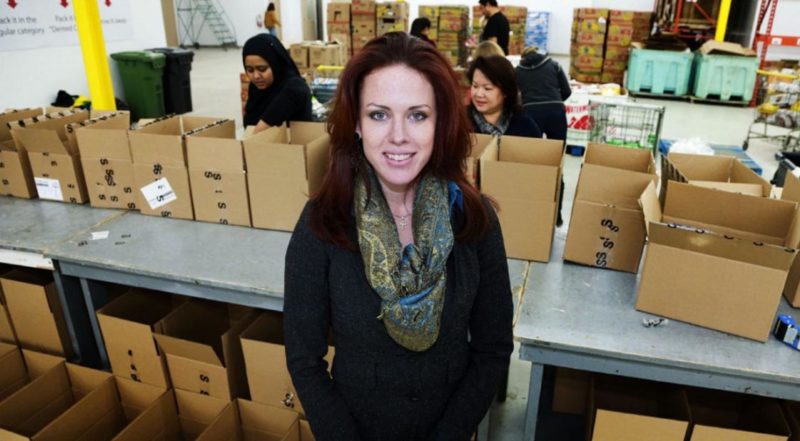 A picture of a smiling woman standing in front of tables loaded with boxes of produce. Many people are in the background