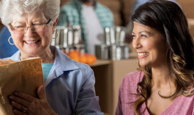 A picture of a smiling woman, next to an older woman, smiling and holding a paper bag with produce