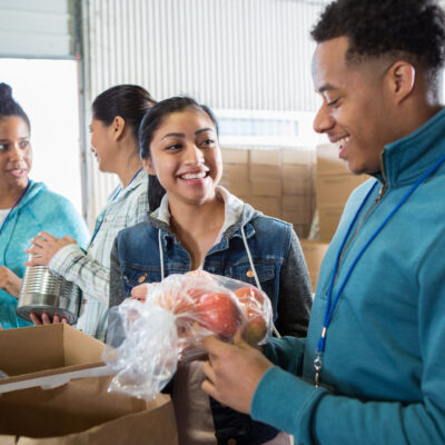 A group of people volunteering at a food bank, smiling and packing food items like canned goods and fresh produce into boxes.