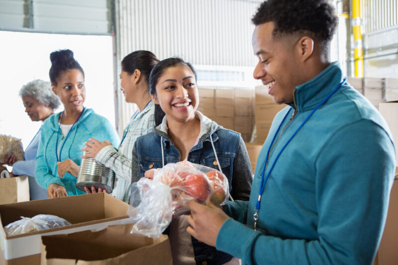 A group of people volunteering at a food bank, smiling and packing food items like canned goods and fresh produce into boxes.