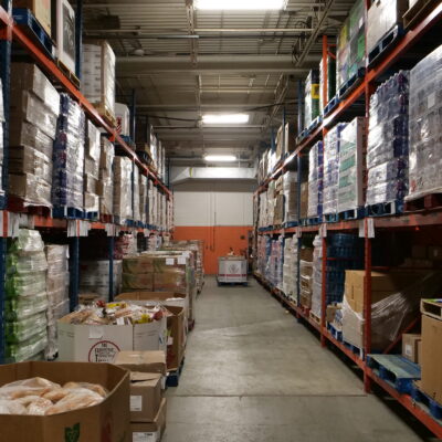 A warehouse aisle with stacks of boxed goods on blue and orange shelves, fostering an environment ripe for collaboration, and a pallet truck in the distance.