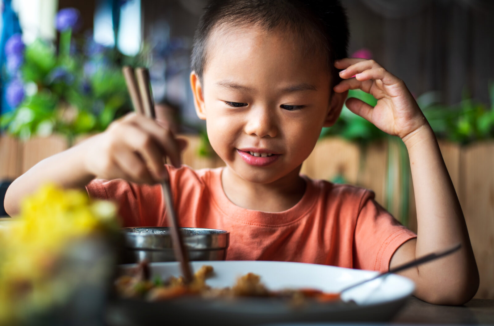 A young boy in an orange shirt uses chopsticks to eat from a bowl at a wooden table, with plants visible in the background.