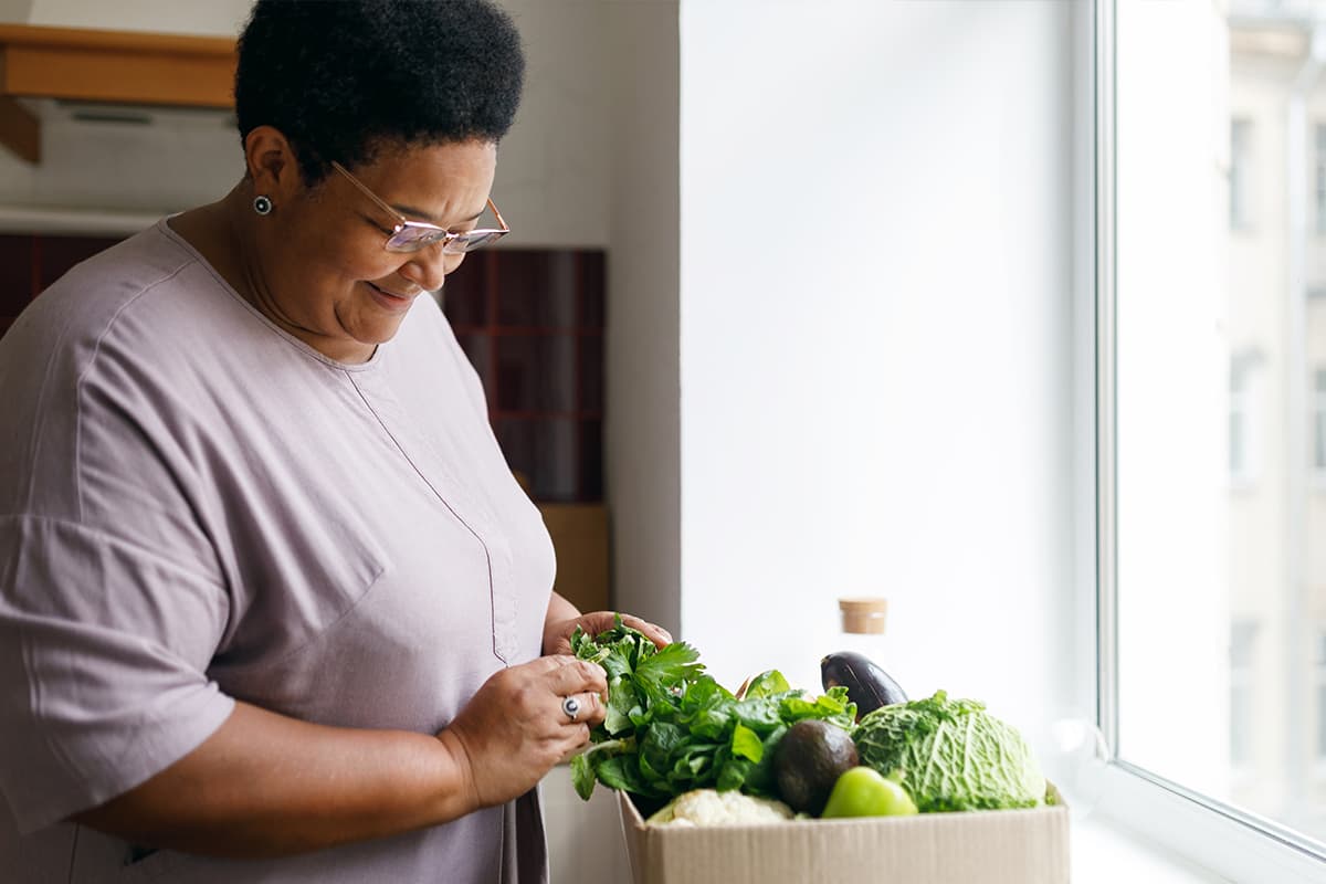 A middle-aged Black woman with a lavender shirt, standing in the kitchen and sorting through a box of fresh green vegetables.