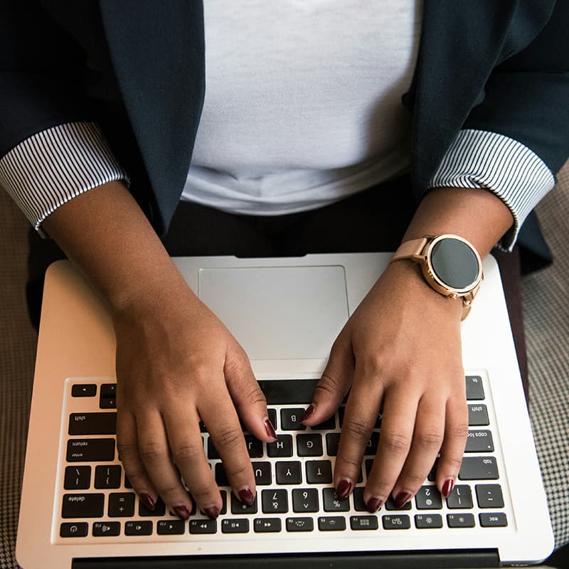 A Black woman wearing a navy suit jacket and a gold watch, typing on a laptop.