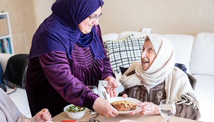 A muslim woman wearing purple handing a plate of fresh food to an older woman, sitting at a kitchen table.