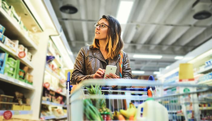 A woman walking through a grocery store aisle, phone in hand, looking in the refrigerator.