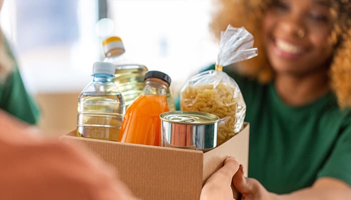 A Black woman in a green shirt handing a box of perishable pasta, cans and juice to another person.