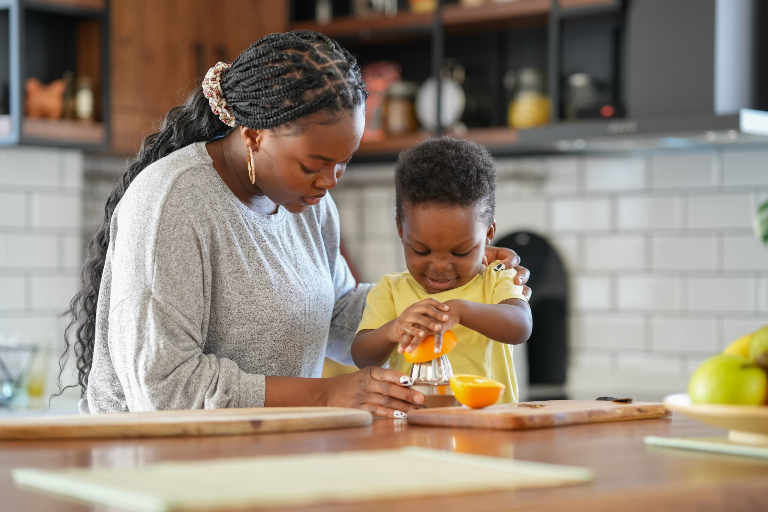 Woman helping a young child squeeze an orange with a juicer in a kitchen. The child is focused on the task, and the woman is smiling supportively.