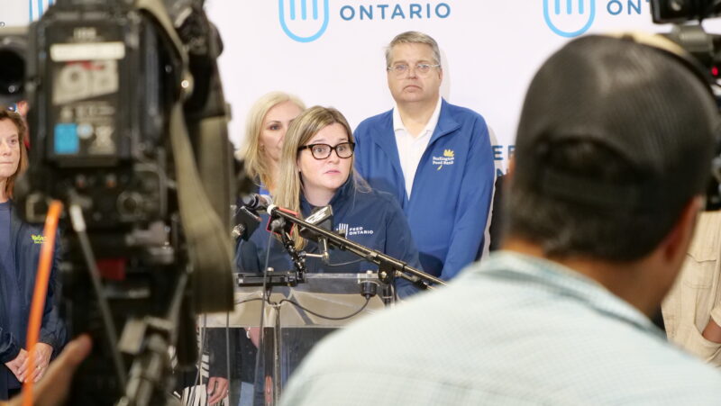 A person speaking at a podium surrounded by microphones, with others standing behind them. Cameras and people are positioned in the foreground.