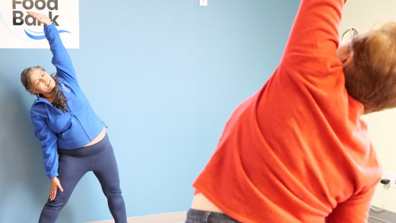 A yoga teacher demonstrating a stretch to a class participant at a food bank.