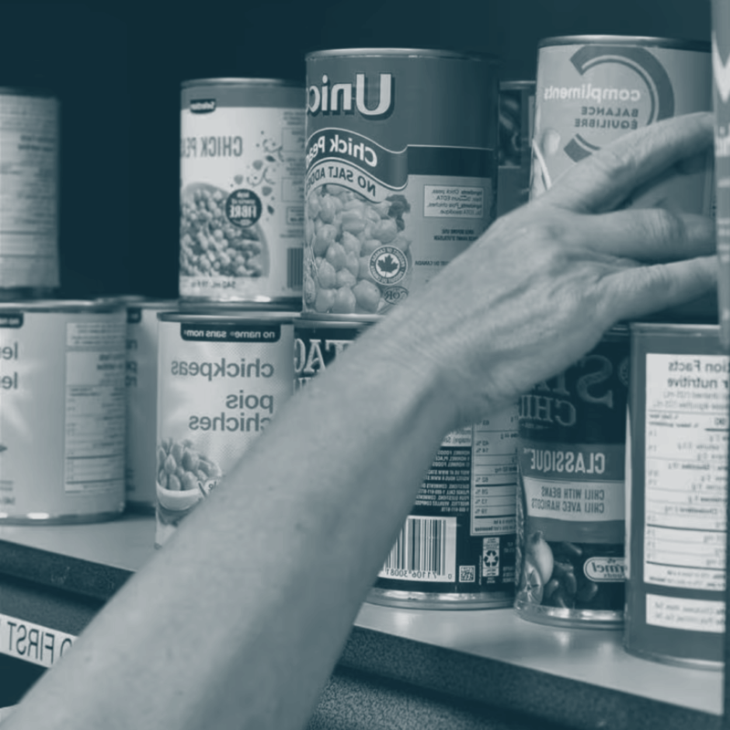 A hand reaches for a can on a food bank shelf.