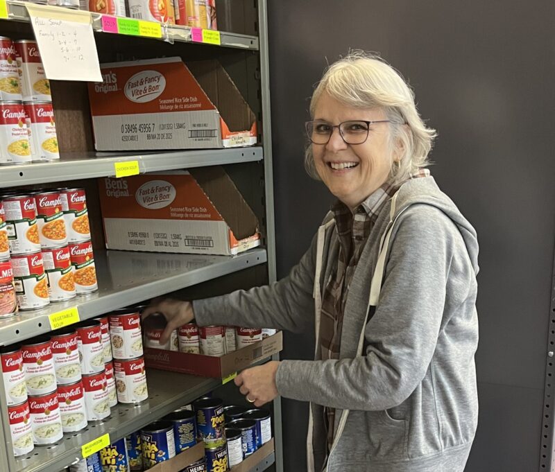 A volunteer smiles standing in front of a shelf at a food bank.
