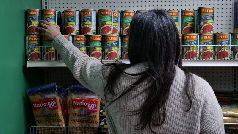 A woman reaches to grab a can of tomato sauce off a shelf at a food bank.