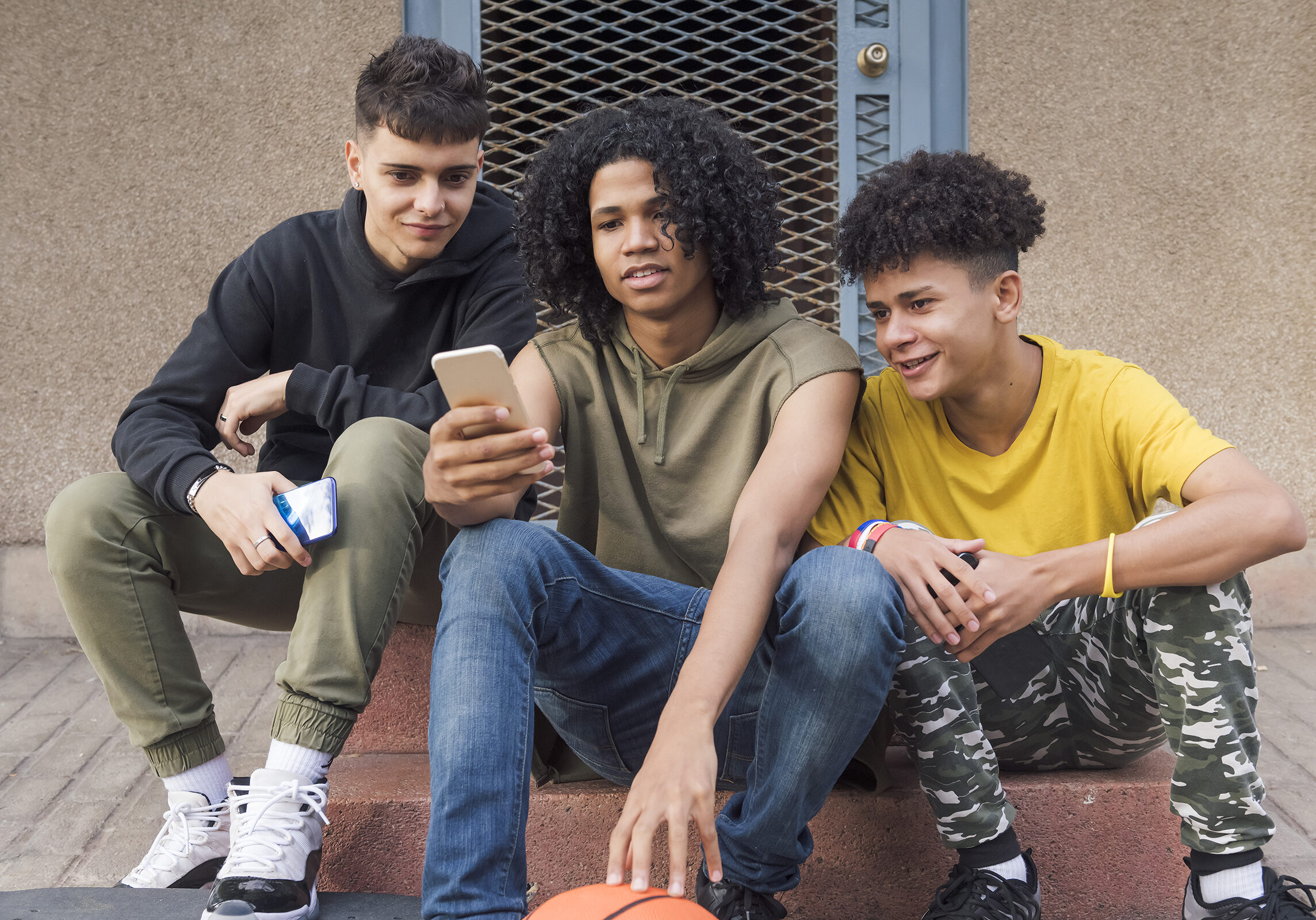 three happy multiethnic teen friends sharing mobile phone sitting together with on street, concept of youth and technology of communication