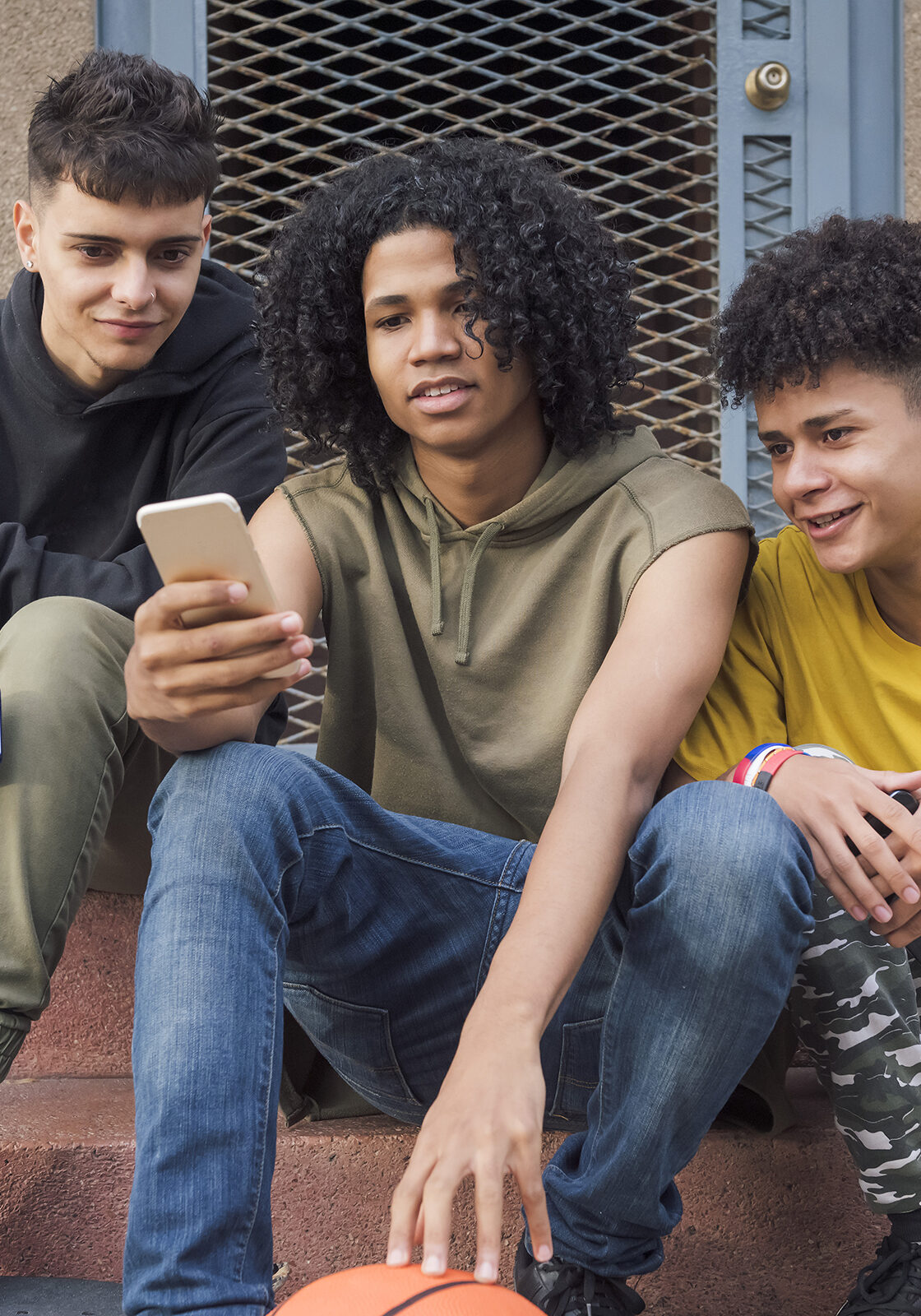 three happy multiethnic teen friends sharing mobile phone sitting together with on street, concept of youth and technology of communication