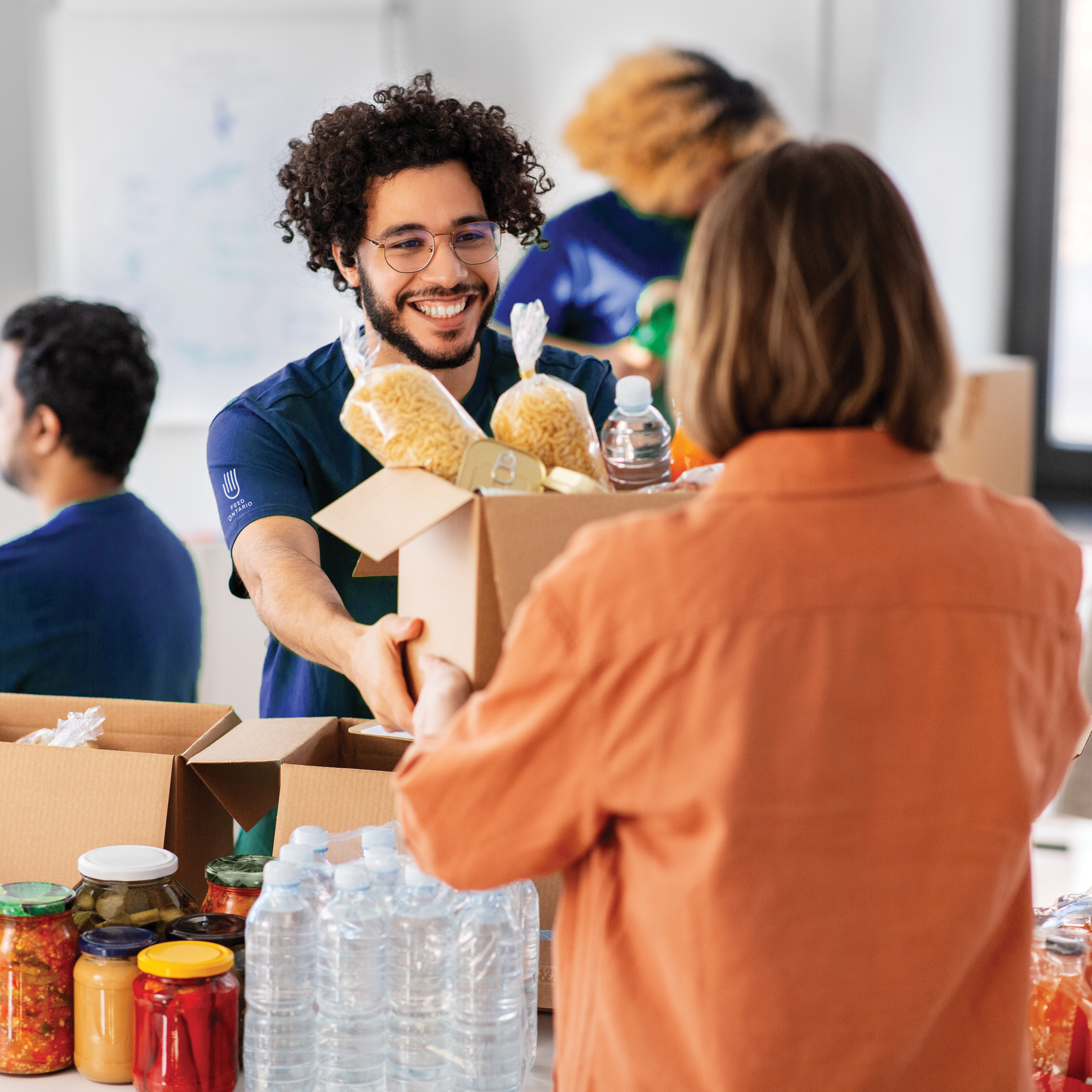 A food banker in a Feed Ontario T-Shirt hands a box of food (pasta and canned goods) to a visitor