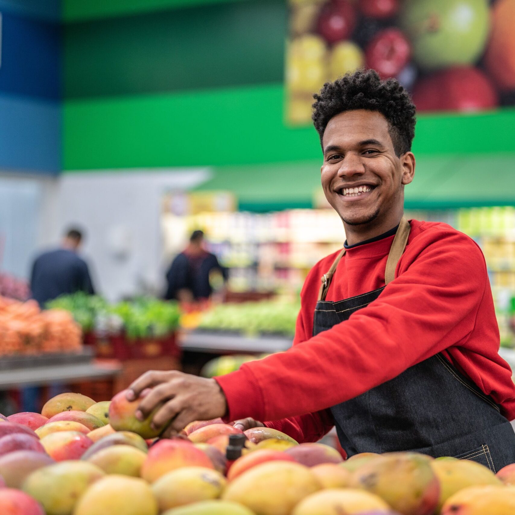 A smiling store employee in a red shirt and black apron arranging fruit in the produce section of a supermarket.