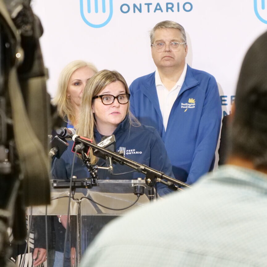 A person speaking at a podium surrounded by microphones, with others standing behind them. Cameras and people are positioned in the foreground.