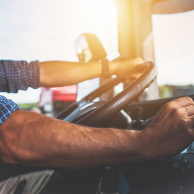 A person wearing a blue checkered shirt is gripping the steering wheel of a vehicle with sunlight streaming through the window.