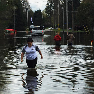 People walk through a flooded street with water up to their knees. A truck and other individuals are visible in the background.