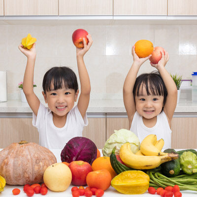 Two children smiling and holding up fruits, surrounded by various fruits and vegetables on a kitchen counter.