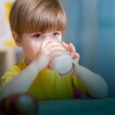 A young child with light brown hair is drinking milk from a glass while wearing a yellow shirt.