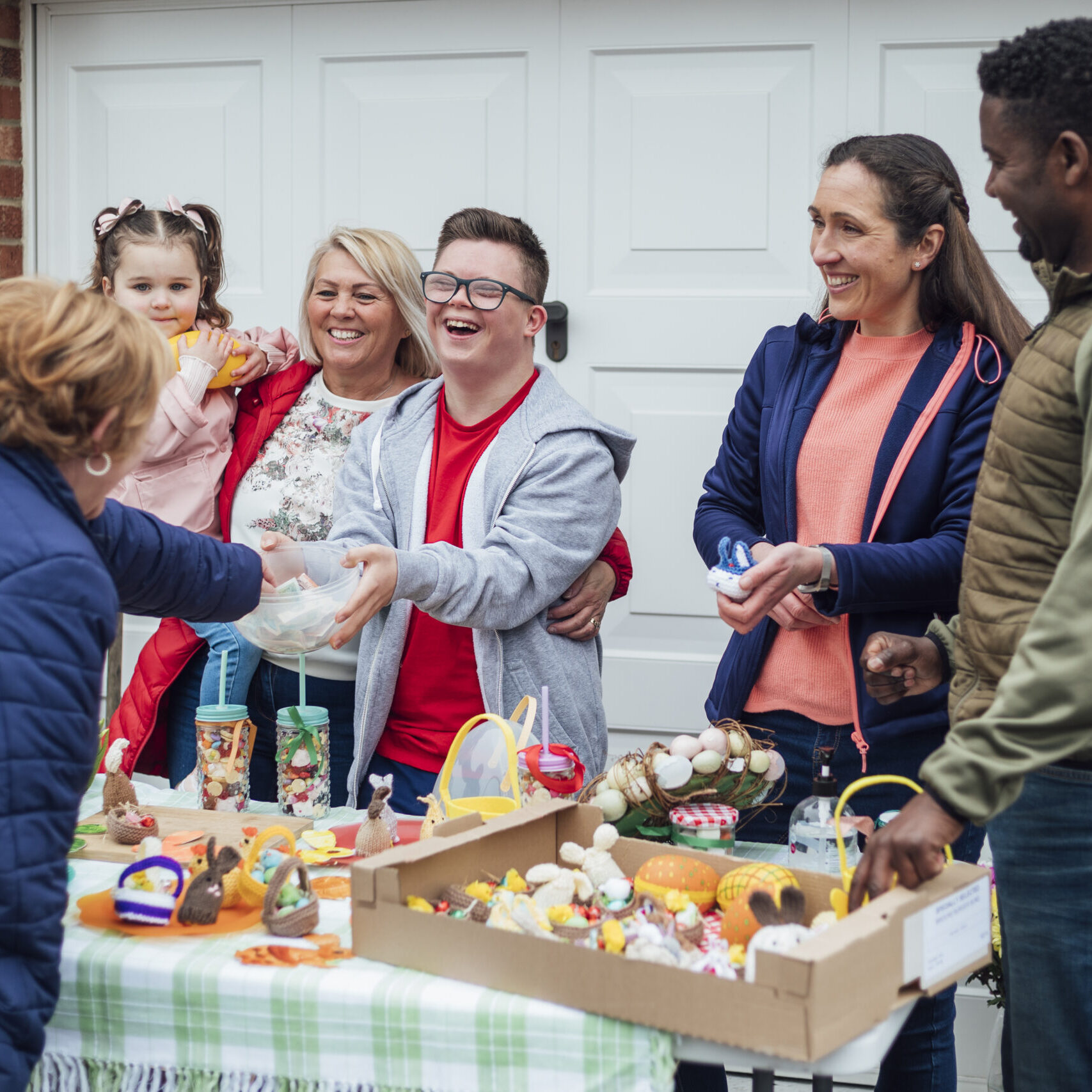 Family having an Easter yard sale in their garden in the North East of England. They have hand made easter crafts on a table and chocolate snacks. One of the sellers has down syndrome and is laughing while collecting money from a customer in a bowl.