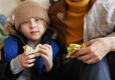 Close up of Caucasian little boy with family in refugee shelter eating sandwich and looking at camera