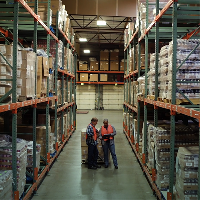 Two workers in an aisle of a large warehouse, one in a high-visibility vest, discussing something while surrounded by shelves stocked with various packages.